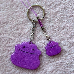 May include: A set of two purple glittery Ditto-shaped keychains. The larger charm is connected to a silver keyring with a braided purple cord and a chain. The smaller charm is attached to the chain. Both charms have a black smiling face.