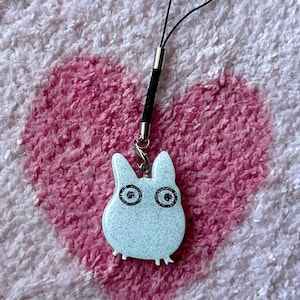 May include: A light blue glittery charm shaped like a cartoon character with large eyes and small feet. It has a black cord and a silver-colored metal clasp. The charm is on a pink heart-shaped background.