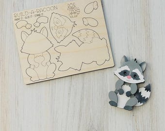 May include: A wooden Build-A-Raccoon puzzle with the text "BUILD-A-RACOON" and instructions "PAINT IT - GLUE IT - STAND IT". The puzzle includes raccoon body parts and a completed raccoon figure in gray, white, and black.