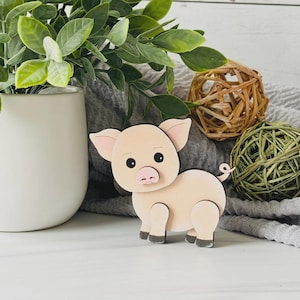 May include: A wooden pig figurine with a light pink body, black eyes, and a pink snout. The pig has black hooves and a curled tail. The figurine is set against a backdrop of greenery and decorative spheres.