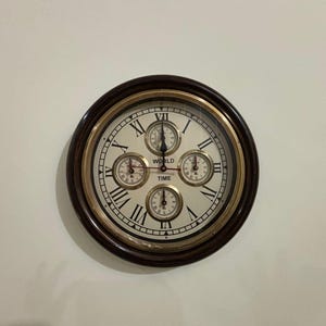 May include: A round wall clock with a dark brown wooden frame and a gold-colored inner ring. The clock face is white with black Roman numerals and four smaller dials, with the words "WORLD TIME" printed on the face.