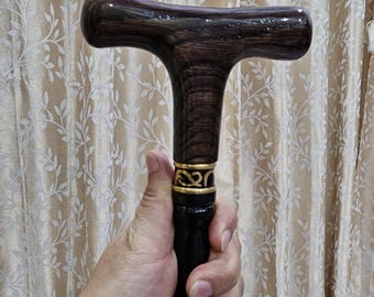 May include: A dark brown wooden cane with a T-shaped handle. The handle and top portion of the cane have a glossy finish. A gold-colored band with an ornate design accents the top of the black shaft.
