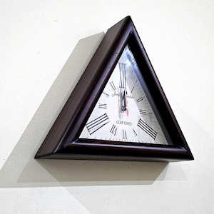 Peut inclure: Une horloge murale triangulaire avec un cadre en bois brun foncé. Le cadran est blanc avec des chiffres romains noirs et le mot "OXFORD" en bas. Les aiguilles sont noires et rouges.