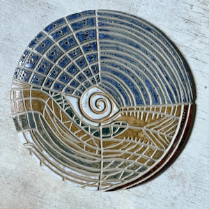 May include: A decorative ceramic plate with a distinctive design. The top half is blue and white with a grid pattern, the bottom half is brown and white with a mosaic design. A white spiral is in the centre. The plate is round and textured.