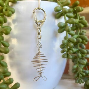 May include: A silver-colored fish-shaped keychain. The fish design is made of wire, with a spiral body and tail fins. The keychain has a clasp for attaching to keys or a bag. The background includes a white pot and green plants.