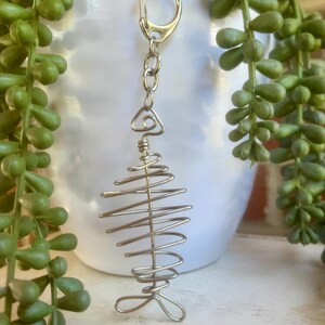 May include: A silver-colored metal fish skeleton keychain. The fish skeleton is made of coiled wire and attached to a chain and a clasp. The background includes a white pot and green plants.