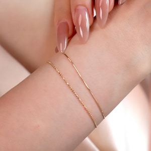 May include: Two delicate gold bracelets adorn a wrist. One bracelet features a fine chain, while the other has a simple, smooth design. The skin tone is light, and the background is a soft, neutral color, highlighting the jewelry.