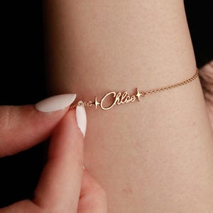 May include: Gold bracelet with the name "Chloe" in cursive. Small star details flank the name. The fine chain is held by a hand with white nail polish. A delicate piece of jewellery.