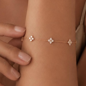 Könnte beinhalten: Ein zartes Armband aus einer goldenen Kette mit drei gleichmäßig verteilten blumenförmigen Anhängern, die jeweils mit vier kleinen, klaren, runden Steinen besetzt sind. Das Armband besteht aus einer feinen Metallgliederkette.