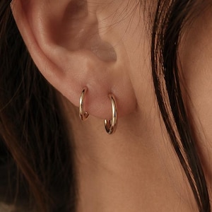 May include: Two small, polished gold hoop earrings worn in the earlobe. The earrings feature a simple, circular design with a smooth, reflective metallic finish. The hoops are of slightly different sizes, with one positioned higher than the other.