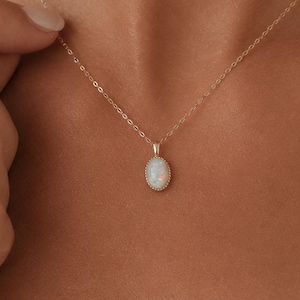 May include: A delicate gold chain necklace featuring a small, oval-shaped opal pendant. The opal displays iridescent flashes of blue and white light, set within a fine, textured gold bezel frame that hangs centered on the thin, polished gold link chain.
