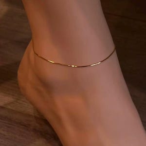 Op de afbeelding: Een delicate goudkleurige enkelband. De dunne ketting wordt om de enkel gedragen. De enkelband is een eenvoudig, elegant sieraad. De achtergrond is een houten oppervlak.