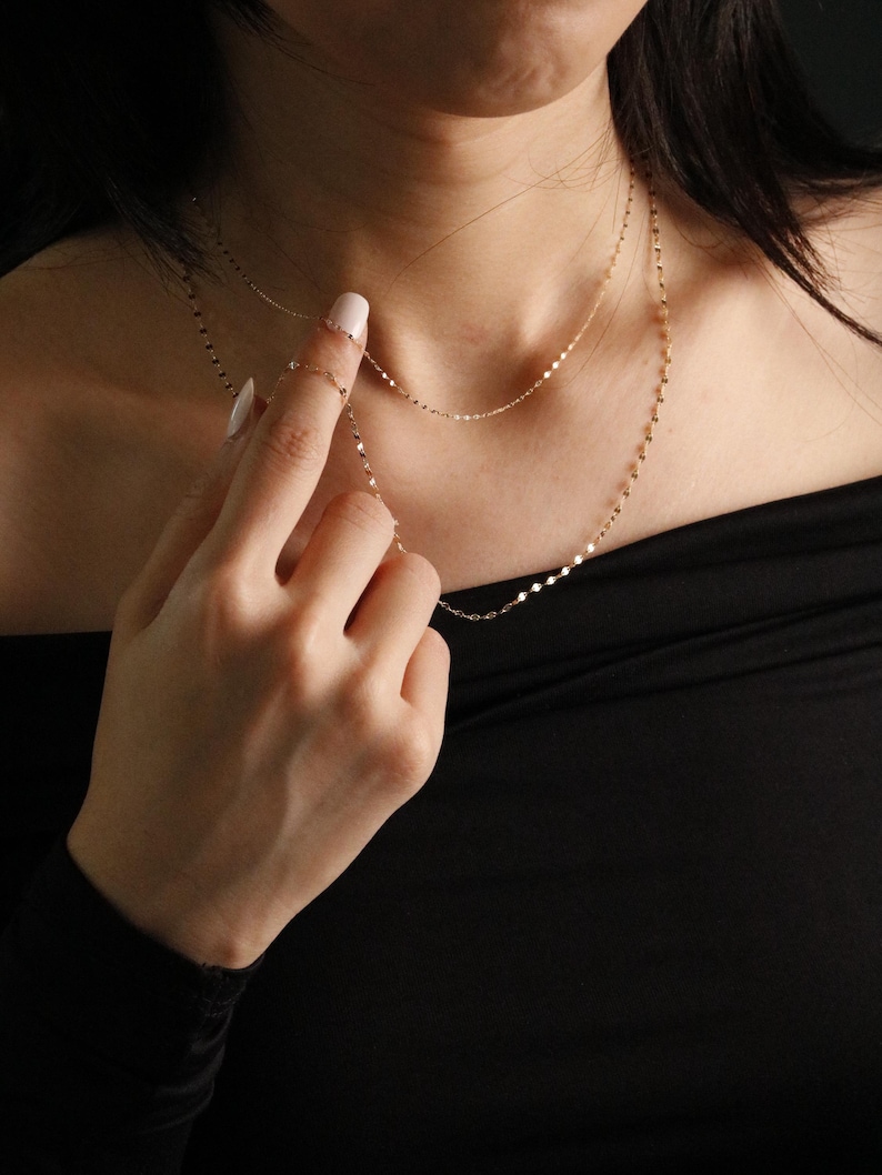 Pu&ograve; includere: Delicate collane d'oro con un design a strati. Le collane presentano piccole maglie rotonde e rettangolari. I gioielli sono indossati su un top nero. L'immagine mostra una mano che tocca una delle collane.