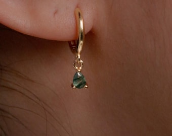 May include: A gold hoop earring featuring a small, teardrop-shaped green gemstone charm. The polished gold hoop is circular and secures through the earlobe, while the dark green, translucent stone hangs from a small gold jump ring at the base.