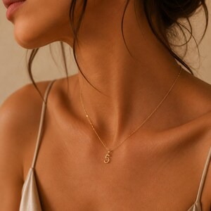 May include: A delicate gold chain necklace featuring a small, minimalist pendant in the shape of the number five. The necklace has a thin, polished gold link chain that rests against the collarbone, showcasing a simple and refined design.