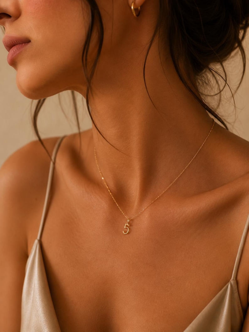 May include: A delicate gold chain necklace featuring a small, minimalist pendant in the shape of the number five. The necklace has a thin, polished gold link chain that rests against the collarbone, showcasing a simple and refined design.