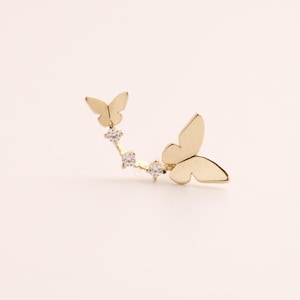 Op de afbeelding: Een gouden oormanchet met twee vlindermotieven en drie heldere, ronde edelstenen. De vlinders bevinden zich aan weerszijden van een gebogen staaf, met de edelstenen langs de boog. De sieraden zijn geplaatst tegen een lichte achtergrond.