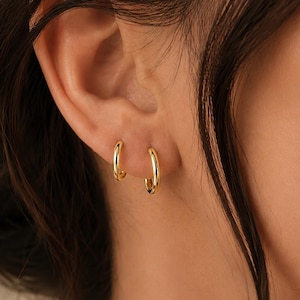 Peut inclure: Deux petites créoles dorées polies portées au lobe de l'oreille. Les boucles d'oreilles présentent une surface métallique lisse et arrondie avec une finition réfléchissante. Chaque anneau est circulaire et d'une épaisseur constante.
