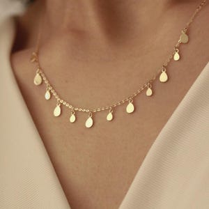 Op de afbeelding: Een delicate gouden ketting met een reeks kleine, druppelvormige hangers. De ketting wordt getoond tegen een zachte, neutrale achtergrond, die het elegante ontwerp van de sieraden benadrukt. De druppels zijn gelijkmatig verdeeld over de ketting.