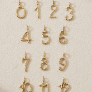 May include: A collection of gold-toned metal number charms ranging from zero to twelve. Each charm features a unique, organic, and slightly irregular wire-like shape with a small loop at the top for attachment, presented in a grid layout.