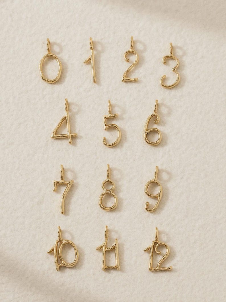 May include: A collection of gold-toned metal number charms ranging from zero to twelve. Each charm features a unique, organic, and slightly irregular wire-like shape with a small loop at the top for attachment, presented in a grid layout.