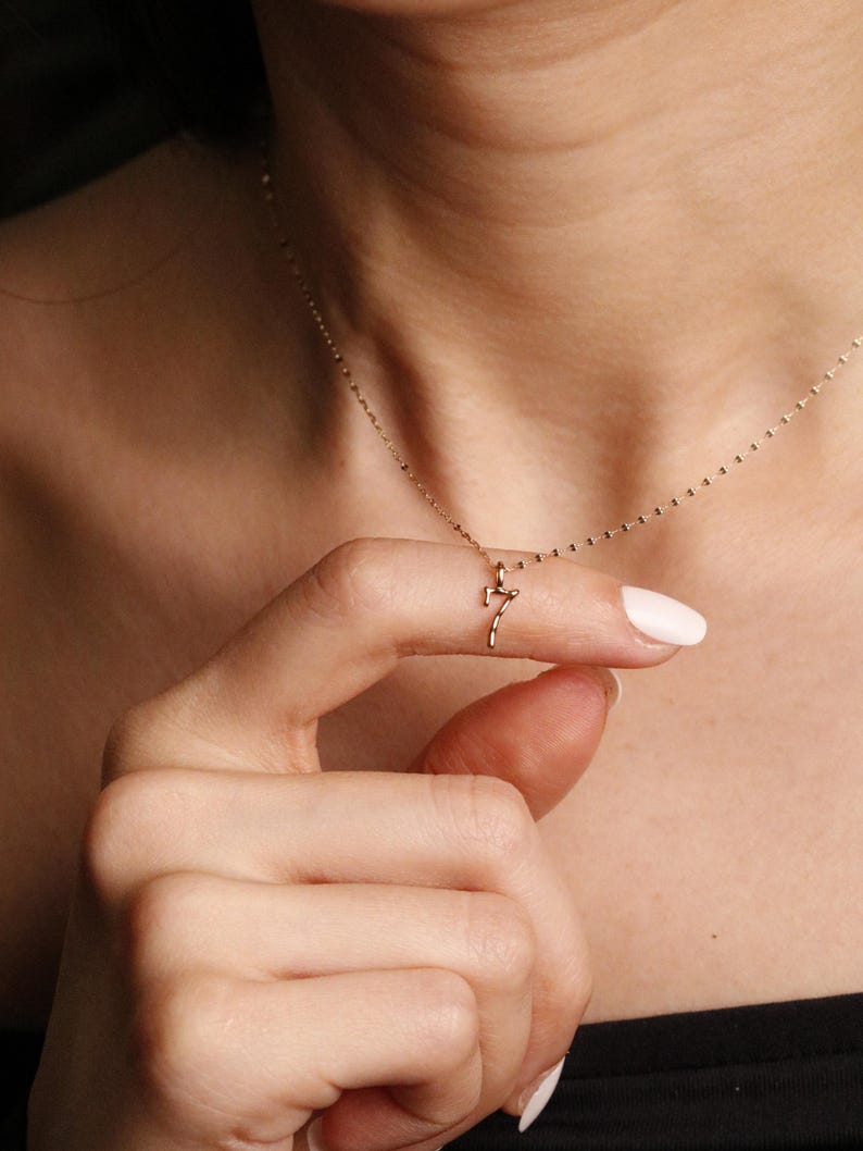 May include: A delicate gold-toned necklace featuring a small, minimalist pendant shaped like the number seven. The pendant hangs from a thin, fine-link chain that rests against the collarbone, showcasing a simple and refined design aesthetic.