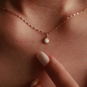Può includere: Una delicata collana d'oro con un piccolo pendente rotondo in opale. Il pendente è incastonato in una lunetta d'oro e pende da una sottile catena. La collana è mostrata sul collo di una persona.