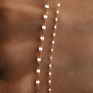 Pu&ograve; includere: Primo piano di due delicate collane d'oro. Ogni collana presenta un motivo ripetuto di piccole maglie ovali allungate. Le catene sono esposte su uno sfondo color carne, evidenziando la sottile lucentezza e il design dei gioielli.