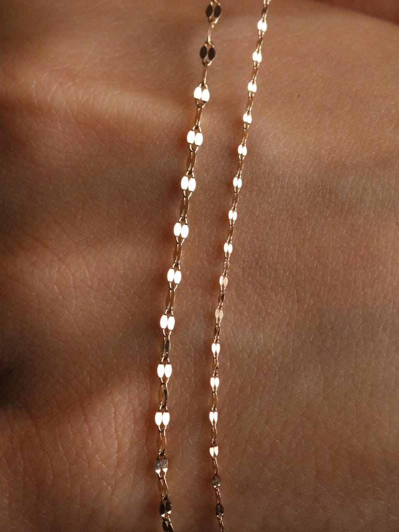 Pu&ograve; includere: Primo piano di due delicate collane d'oro. Ogni collana presenta un motivo ripetuto di piccole maglie ovali allungate. Le catene sono esposte su uno sfondo color carne, evidenziando la sottile lucentezza e il design dei gioielli.
