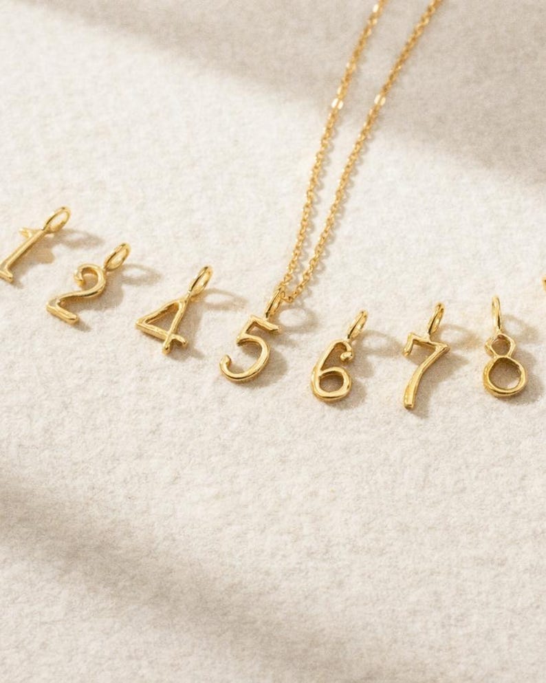 May include: A gold-toned necklace featuring a number five pendant, accompanied by a collection of individual gold-toned number charms ranging from one to eight, each equipped with a small loop for attachment to a chain or jewelry component.