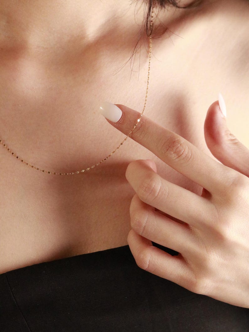 Close-up of a delicate 14k solid gold chain necklace with small reflective links, shown resting on skin and fingers to show its fine, lightweight design