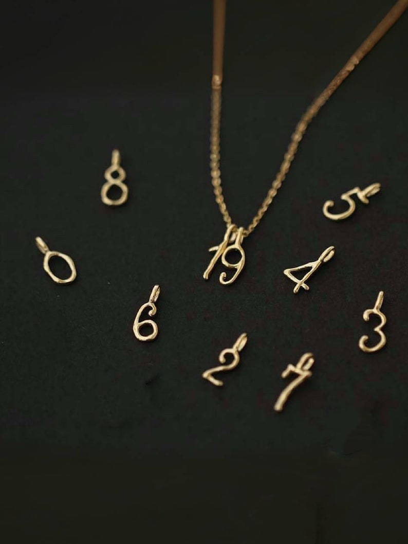 May include: A gold-toned necklace chain with the numbers 1 and 9 attached as pendants, surrounded by individual gold-toned number charms including 0, 2, 3, 4, 5, 6, 7, and 8, each featuring a small loop at the top for attachment to a chain.