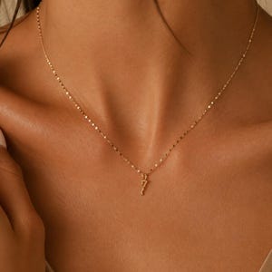 May include: A delicate gold chain necklace featuring a small, slender pendant shaped like the number seven. The necklace has a fine, reflective link design that catches the light, resting against the collarbone with a simple, minimalist aesthetic.