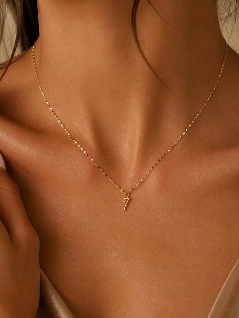 May include: A delicate gold chain necklace featuring a small, slender pendant shaped like the number seven. The necklace has a fine, reflective link design that catches the light, resting against the collarbone with a simple, minimalist aesthetic.