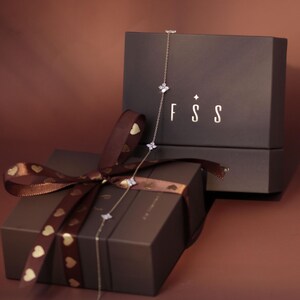 May include: A delicate silver-toned chain necklace featuring small, four-petaled flower-shaped charms with clear stones. The necklace is draped over two dark brown gift boxes, one tied with a brown ribbon featuring gold heart patterns.