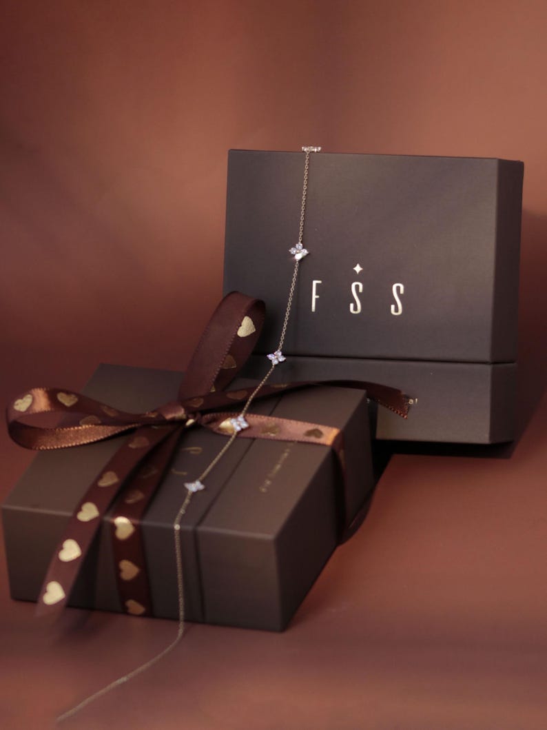 May include: A delicate silver-toned chain necklace featuring small, four-petaled flower-shaped charms with clear stones. The necklace is draped over two dark brown gift boxes, one tied with a brown ribbon featuring gold heart patterns.