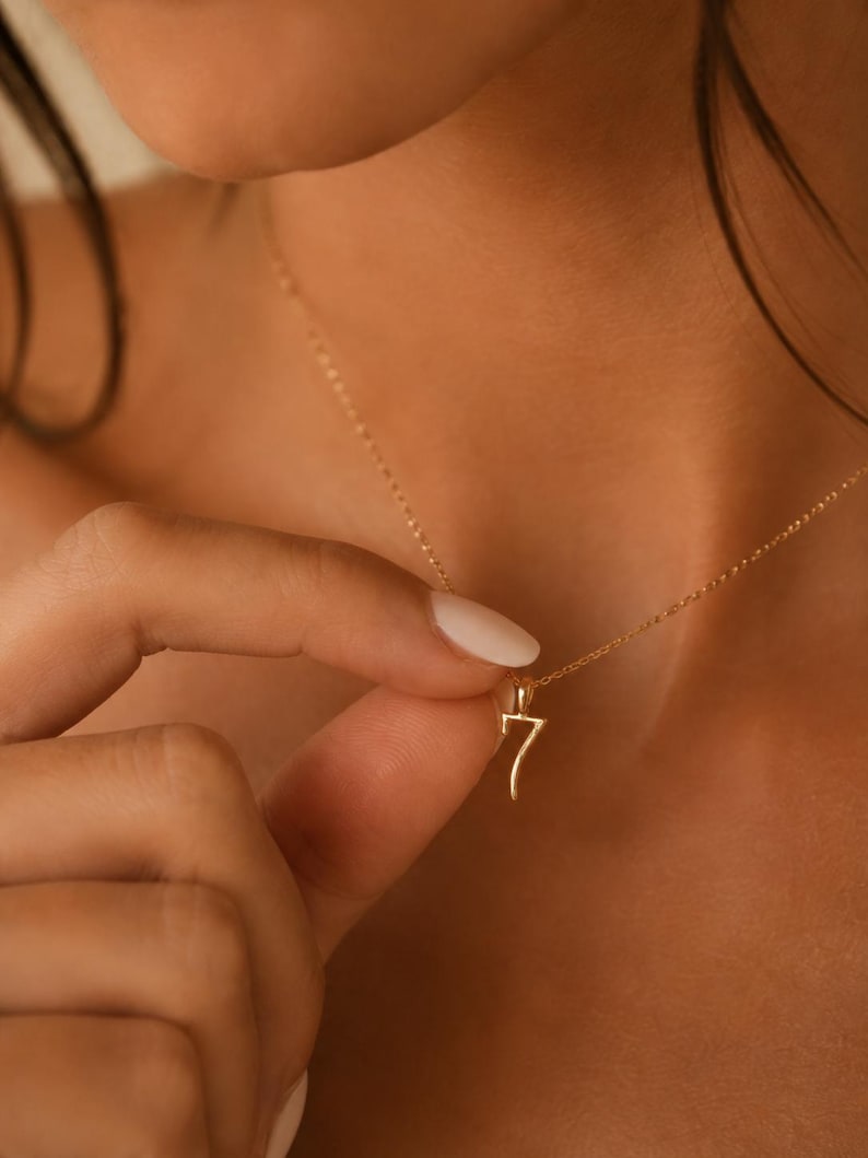 May include: A gold-toned necklace featuring a delicate chain and a pendant shaped like the number seven. The pendant has a smooth, polished finish and is suspended from a thin, matching gold-toned link chain that rests against the collarbone area.