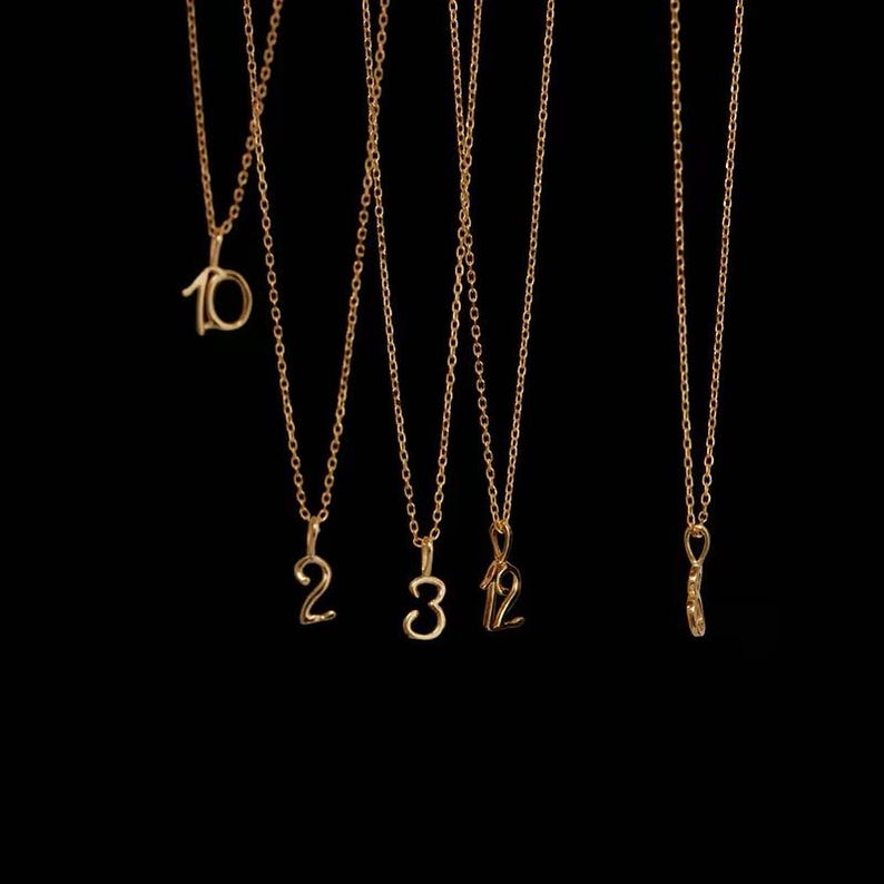 May include: Five gold-toned necklaces hanging vertically against a black background. Each necklace features a delicate chain and a pendant shaped as a numeral. The pendants display the numbers 10, 2, 3, 12, and a single digit that is partially visible.