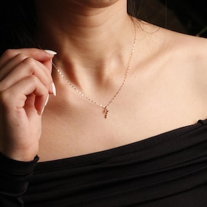 May include: A delicate gold chain necklace featuring a small, slender pendant. The chain is composed of tiny, flat, reflective gold links that catch the light, creating a shimmering effect around the neck. The pendant is a simple, thin gold shape.