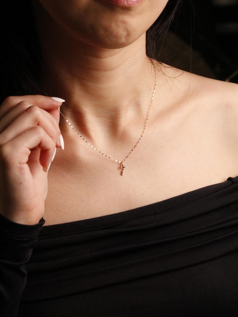 May include: A delicate gold chain necklace featuring a small, slender pendant. The chain is composed of tiny, flat, reflective gold links that catch the light, creating a shimmering effect around the neck. The pendant is a simple, thin gold shape.