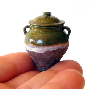 Może przedstawiać: Miniaturowy ceramiczny garnek z pokrywką i dwoma małymi uchwytami. Górna część wykończona jest błyszczącym oliwkowozielonym szkliwem, natomiast dolna sekcja ma teksturowane, ciemnobrązowe matowe wykończenie, oddzielone cienkim, białym pasem.
