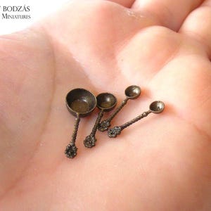 Op de afbeelding: Een set van vier miniatuur bronskleurige metalen maatlepels met ronde kommen en gedraaide handvatten met decoratieve bloemachtige uiteinden. De lepels zijn gerangschikt in aflopende grootte en vertonen een metallic afwerking.