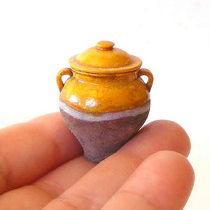 Może przedstawiać: Miniaturowy ceramiczny garnek z pokrywką i dwoma bocznymi uchwytami. Górna część jest szkliwiona na błyszczący musztardowy kolor, podczas gdy dolna sekcja ma matowe, teksturowane wykończenie w kolorze ciemnego brązu. Tekst na górze brzmi ERZSÉBET BODZÁS HUNGARIAN MINIATURES.