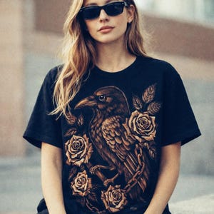 Puede incluir: Camiseta negra con una ilustración detallada de un cuervo posado en una rama con rosas. El cuervo y las rosas son en tonos marrones y dorados. La camiseta tiene mangas cortas y cuello redondo.