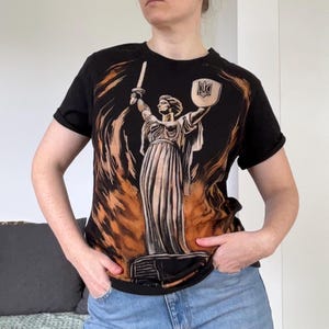 Könnte beinhalten: Schwarzes T-Shirt mit einer Grafik einer Statue, die ein Schwert und einen Schild vor einem feurigen Hintergrund hält. Der Schild hat ein Wappen. Das T-Shirt hat kurze Ärmel.