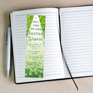Pode incluir: Um marcador de página com o texto "I WILL FIND MY OWN Sacred Grove" em verde e branco. O marcador de página tem um design de folhas verdes e está colocado dentro de um caderno aberto com uma caneta.