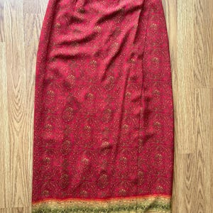 May include: A red wrap skirt with a floral pattern in gold and brown. The skirt has a button closure and a decorative border at the hemline with green, yellow, and orange stripes. The skirt is made of a lightweight fabric.