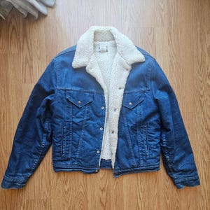 May include: A blue denim jacket with a cream-colored, fluffy shearling lining and collar. The jacket has two chest pockets with button closures and two side pockets. The jacket is buttoned up and is lying on a wooden surface.