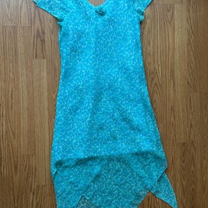 May include: A turquoise dress with a V-neck and short sleeves. The dress features a subtle, all-over pattern and a unique asymmetrical hemline. A small floral detail accents the neckline. The dress is made of a lightweight, flowing fabric.