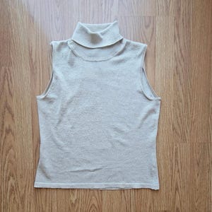 May include: A sleeveless, light beige turtleneck sweater. The sweater is made of a soft, textured knit fabric. The turtleneck collar is folded over. The sweater is laying on a wooden surface.